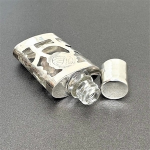 Puede incluir: Una botella de plata y vidrio con un diseño decorativo y un tapón de plata separado. La botella está abierta y el tapón está acostado a su lado.