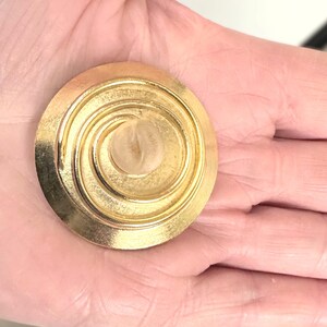 Puede incluir: Un broche circular dorado con un diseño de espiral y un centro de vidrio transparente.