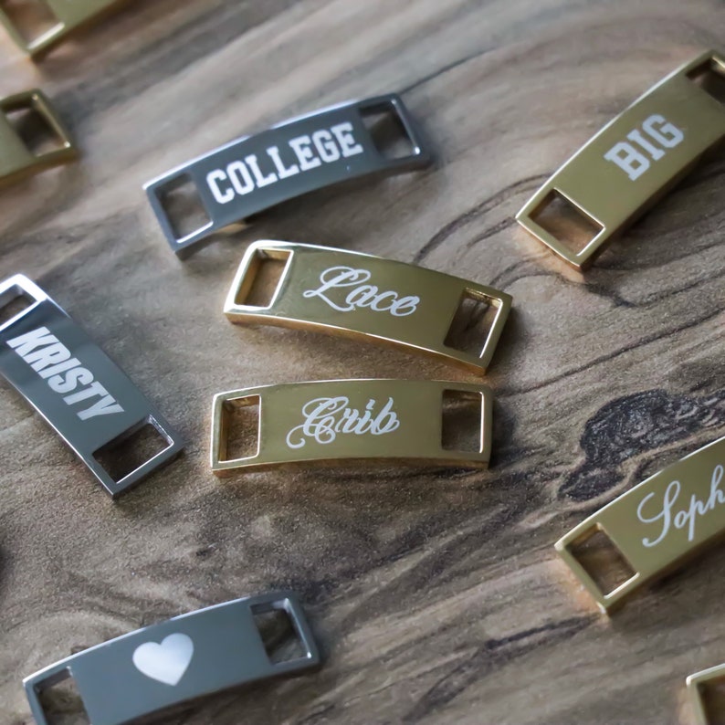 May include: A collection of gold and silver shoe charms with various engraved words, including "College", "Lace", "Crib", "Big", "Kristy", and "Soph". One charm features a heart design.