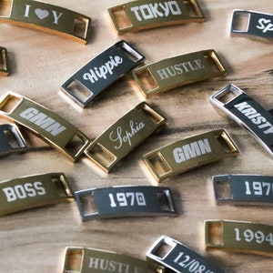 May include: A collection of gold and silver metal shoe charms with various words and numbers engraved on them, including "GMN", "Sophia", "Hippie", "Hustle", "Tokyo", "Boss", "1970", "1994", "Kristy", and "I &hearts; Y".