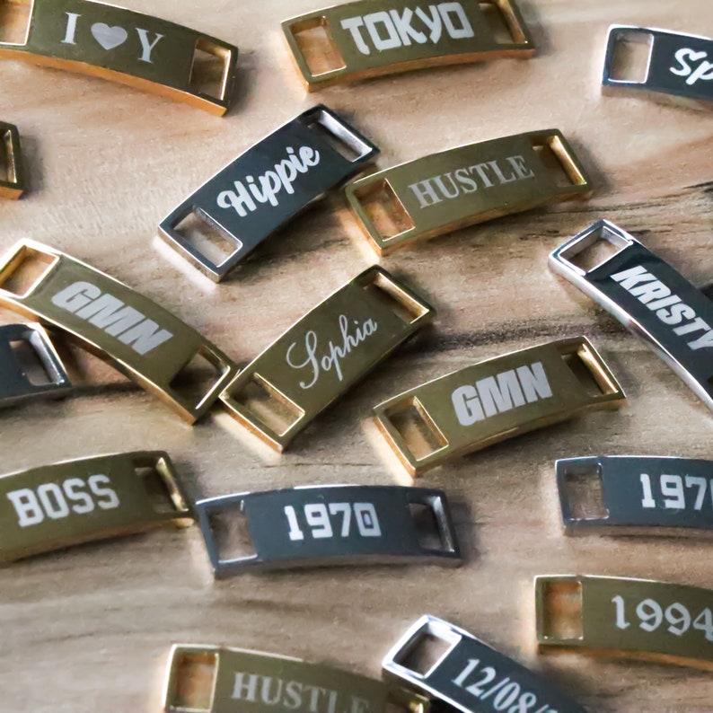 May include: A collection of gold and silver metal shoe charms with various words and numbers engraved on them, including "GMN", "Sophia", "Hippie", "Hustle", "Tokyo", "Boss", "1970", "1994", "Kristy", and "I &hearts; Y".