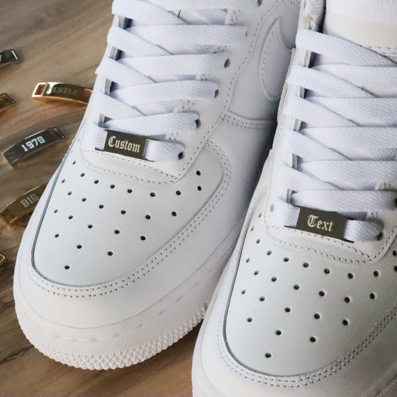 May include: White leather sneakers with white laces and silver metal lace locks. The lace locks have the words "Custom" and "Text" engraved on them.