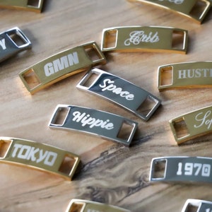 May include: A collection of gold and silver metal shoe charms with various words engraved on them, including "GMN", "Space", "Hippie", "Tokyo", "Cribb", "Hustle", and "1970".