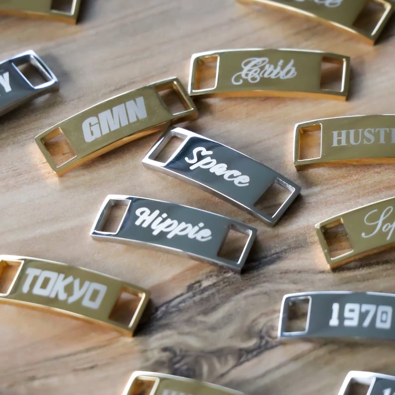 May include: A collection of gold and silver metal shoe charms with various words engraved on them, including "GMN", "Space", "Hippie", "Tokyo", "Cribb", "Hustle", and "1970".