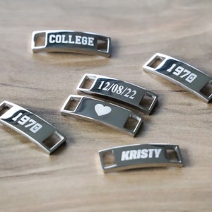 May include: Five silver shoe charms with different engravings. The charms read "COLLEGE", "1970", "12/08/22" with a heart, "1970", and "KRISTY".