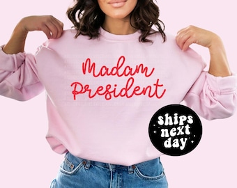 May include: Pink sweatshirt with the text "Madam President" in red lettering. The sweatshirt has a black circle with white text that says "ships next day".