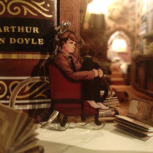 Könnte beinhalten: Ein klarer Acryl-Schlüsselanhänger mit einer Cartoon-Illustration einer Person in einem Sherlock-Holmes-Outfit, die in einem roten Stuhl sitzt. Der Schlüsselanhänger befindet sich vor einem großen Buch mit dem Text "SIR ARTHUR CONAN DOYLE".