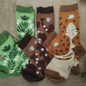 Könnte beinhalten: Verschiedene Socken mit Kaffee- und Teemotiven. Designs umfassen Teekannen, Blumenmuster, Kaffeetassen und Kaffeebohnenmotive. Die Socken sind in Grün-, Braun- und Beigetönen gehalten.