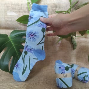 Può includere: Calzini azzurri con un motivo floreale di fiori blu e viola e steli verdi. Un calzino è tenuto in mano, mentre vengono mostrate due paia arrotolate. I calzini hanno un polsino azzurro e sono su una superficie di juta.