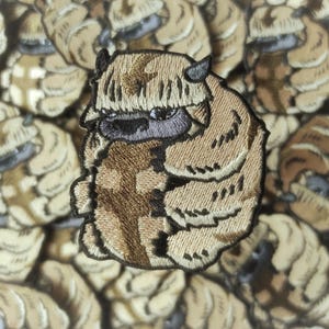 Könnte beinhalten: Gestickter Aufnäher mit einer cartoonartigen, bisonähnlichen Kreatur mit beigefarbenem und braunem Fell, schwarzen Hörnern und einem violetten Gesicht. Der Aufnäher ist von einem Hintergrund aus ähnlichen Aufnähern umgeben. Für Animationsfans.