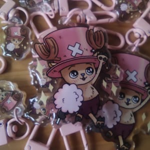 Op de afbeelding: Roze acryl sleutelhangers met een stripfiguur met een roze hoed en bruine geweien. Het personage draagt een paarse outfit en heeft een witte wolkachtige vorm. De sleutelhangers hebben roze sluitingen.