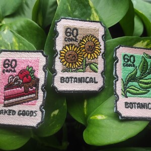 Op de afbeelding: Drie geborduurde patches die op postzegels lijken. Eén toont een stuk cake met bessen en de tekst "BAKED GOODS". Een andere toont zonnebloemen met "BOTANICAL". De laatste toont bladeren met "BOTANICAL". Elke patch heeft "60 cent".