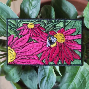 Pode incluir: Patch bordado com um design vibrante de flores de cone rosa com centros amarelos e uma abelha. O patch tem um fundo verde e uma borda preta, perfeito para adicionar a roupas ou acessórios.