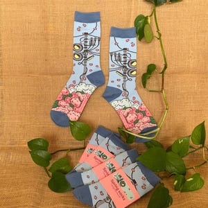 Könnte beinhalten: Zwei Paar hellblaue Socken mit floralem und mechanischem Design. Die Socken haben ein rosa-weißes Blumenmuster an der Spitze und eine mechanische Illustration am Bein. Mehrere gefaltete Sockenpaare sind ebenfalls sichtbar.