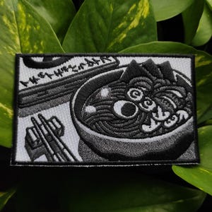 Puede incluir: Parche bordado en blanco y negro que representa un cuenco de ramen con palillos. El parche rectangular incluye texto japonés. El diseño detallado es adecuado para aplicar en ropa o accesorios. Un artículo con temática de comida.
