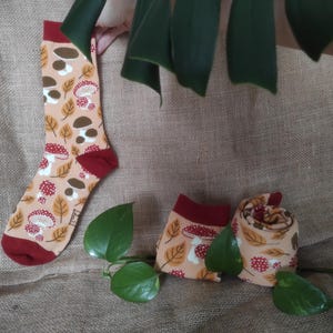 Könnte beinhalten: Beige Socken mit einem Pilz- und Blättermuster, mit roten Akzenten an Zehen und Bündchen. Zwei weitere Socken sind gefaltet und gerollt und zeigen das gleiche Design. Die Socken werden auf einer Jutefläche präsentiert.