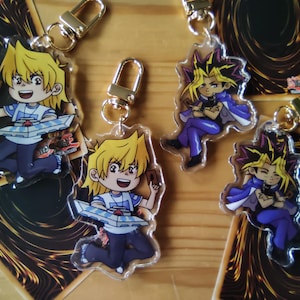 Könnte beinhalten: Acryl-Schlüsselanhänger mit Anime-Charakteren und goldfarbenen Verschlüssen. Die Schlüsselanhänger zeigen zwei verschiedene Charaktere mit stacheligen gelben Haaren und verschiedenen Posen. Die Schlüsselanhänger liegen auf Sammelkartenspielkarten.