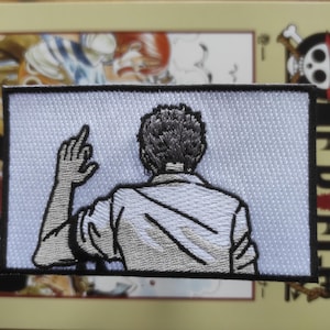 Può includere: Toppa ricamata raffigurante una figura vista da dietro, che alza un dito. La toppa è rettangolare con sfondo bianco e bordo nero. La figura indossa una camicia bianca. La toppa è sopra un libro.