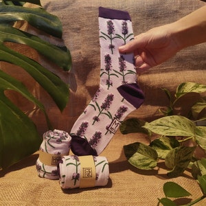 May include: Light purple socks featuring a lavender floral design. The image shows a single sock held up, alongside two rolled pairs. The socks have purple cuffs and heels. The socks are displayed on a textured surface.