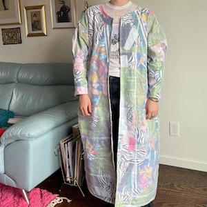 May include: A long, quilted coat with a patchwork design featuring zebra print, floral patterns, and pastel colors. The coat has a collarless design and a loose fit. The coat is worn over a white t-shirt and black jeans.