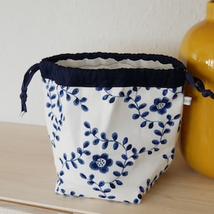 May include: A white drawstring bag with a blue floral pattern and a navy blue drawstring. The bag is sitting on a light wood surface next to a yellow vase.