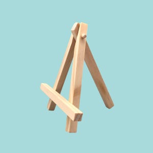 May include: A miniature wooden easel, constructed from light-coloured wood. It has a three-legged structure with a horizontal support. The background is a solid, pale blue.
