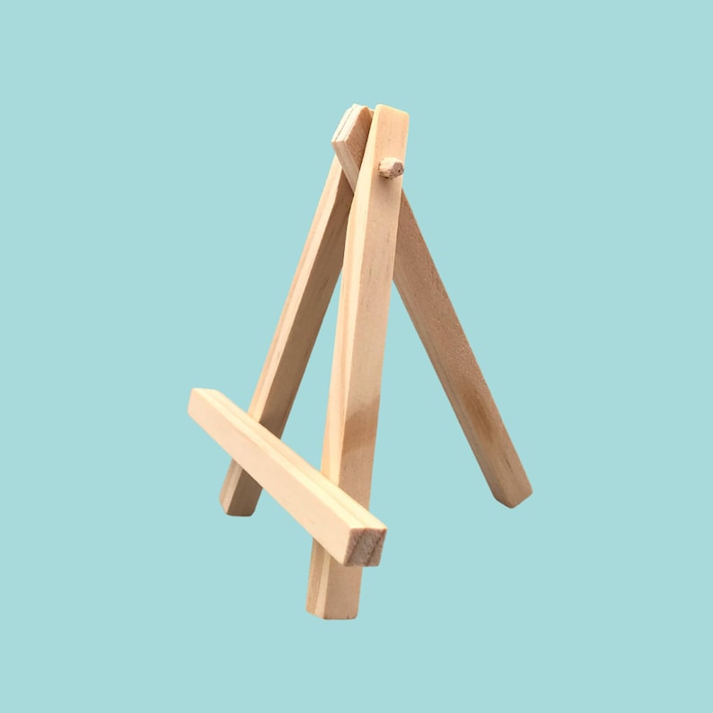 May include: A miniature wooden easel, constructed from light-coloured wood. It has a three-legged structure with a horizontal support. The background is a solid, pale blue.