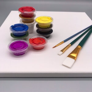 K&ouml;nnte beinhalten: Eine wei&szlig;e Leinwand mit einem Malen-nach-Zahlen-Set. Das Set enth&auml;lt kleine Plastikbeh&auml;lter mit verschiedenen Farben, darunter Rot, Gelb, Blau und Lila. Drei Pinsel mit gr&uuml;nen Griffen und goldenen Zwingen sind ebenfalls enthalten.
