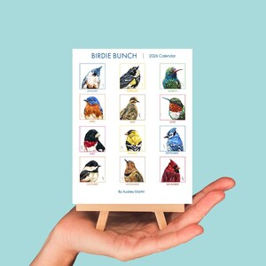May include: A 2026 calendar, "Birdie Bunch", with watercolour illustrations of birds for each month. The calendar is displayed on a small wooden easel. The artist's name, Audrey Martin, is at the bottom of the calendar.