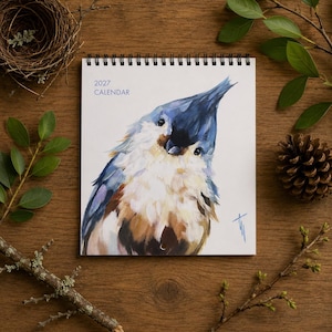 May include: A 2027 calendar with a spiral binding at the top. The cover features a painterly illustration of a tufted titmouse bird with blue, white, and brown feathers. The text 2027 CALENDAR is printed in blue in the upper left corner.