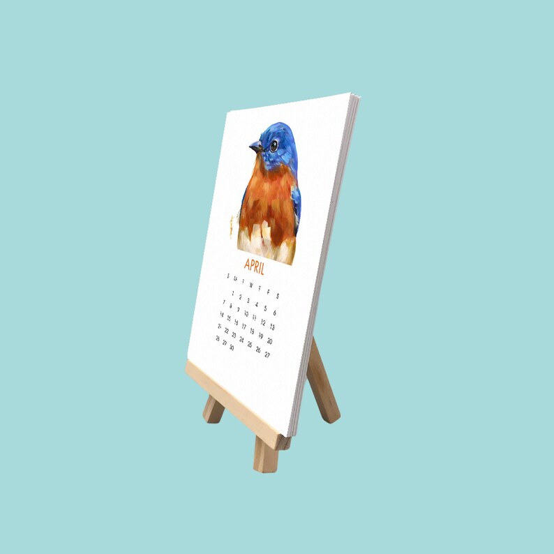 May include: A desk calendar for April, featuring a watercolour painting of a bluebird. The calendar is displayed on a small wooden easel. The bird has blue, orange, and white feathers.