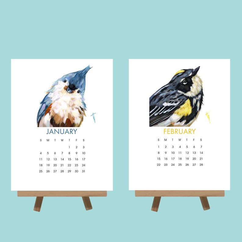 May include: Two small wooden easels each hold a calendar page. The January page features a blue and tan bird, while the February page shows a black, yellow, and white bird. Each calendar page displays the month and days.