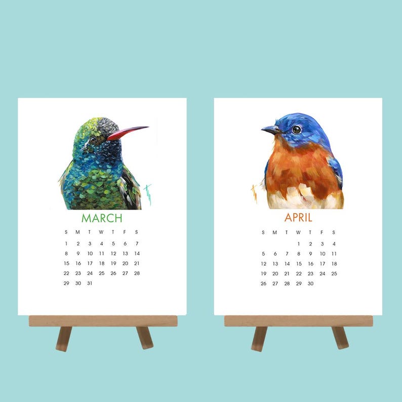 May include: Two small wooden easels display watercolour art calendars. One calendar shows a hummingbird with the month of March, the other a bluebird with the month of April. Each calendar has a corresponding calendar grid.