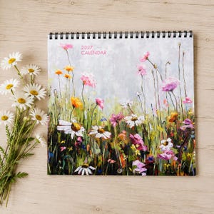 May include: A 2027 wall calendar featuring an impressionistic painting of a wildflower meadow with white, pink, and yellow flowers against a soft grey background. A bouquet of fresh white daisies with yellow centres sits to the left of the calendar.