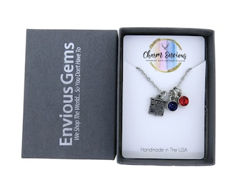 Op de afbeelding: Een zilveren ketting met een New York Rangers-bedel, een blauwe kristal en een rode kristal. De ketting zit in een klein grijs doosje met de woorden "Envious Gems" en "Charm Envious" erop gedrukt.