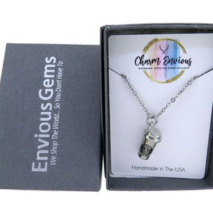 May include: A silver necklace with a charm of a coffee cup. The necklace is in a black box with the text "Envious Gems" and "Charm Envious" on it. The box is on a white background.