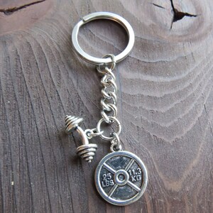 May include: Silver keychain with a dumbbell and a weight plate charm. The weight plate has the text "25 LBS" and "11.3 KG" printed on it.