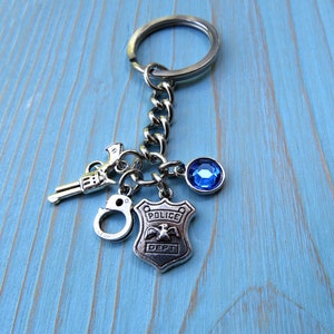 May include: Silver keychain with a police badge charm, handcuffs, a toy gun, and a blue rhinestone. The badge has the words "Police Dept" on it.