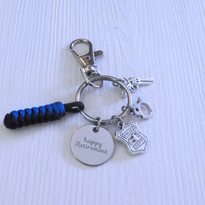 May include: A silver keychain with a blue and black braided cord, a silver charm with the text "happy retirement", a silver police badge charm, a silver handcuff charm, and a silver gun charm.