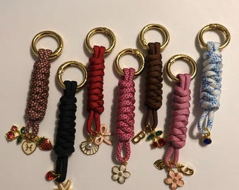Puede incluir: Una colección de ocho llaveros con aros dorados y cordones trenzados en varios colores, incluyendo rosa, rojo, negro y marrón. Cada llavero presenta un dije, como una estrella de mar, una flor o una cereza. El texto "@lovegabbyco" es visible.