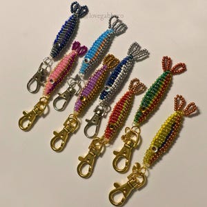 May include: A collection of beaded fish keychains in various colors, including blue, pink, purple, red, green, and yellow. Each keychain features a gold-colored clasp and a beaded tail. The fish have small, bead eyes.