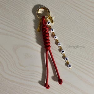 May include: A red paracord keychain with gold accents. The keychain features a braided design and a gold ring. A gold chain with white letter beads spells out a name. A small gold charm and a gold tassel are also attached.