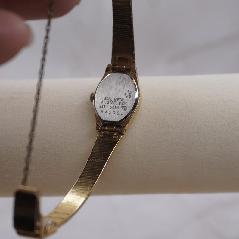 May include: A gold-toned wristwatch with a rectangular face and a delicate, linked band. The watch's back is silver with the text "BASE METAL ST. STEEL BACK" and other markings. The watch is displayed against a neutral background.