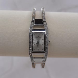 Puede incluir: Reloj Anne Klein plateado con esfera rectangular y correa plateada. La esfera del reloj tiene números romanos y el logotipo de Anne Klein. La correa y los lados de la esfera están incrustados con pequeños cristales transparentes.