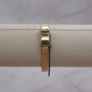 May include: A gold-toned bracelet with a rectangular design. The bracelet features a clasp with a safety chain. The bracelet is displayed on a neutral-colored surface.