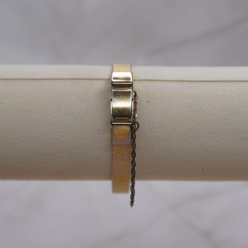 May include: A gold-toned bracelet with a rectangular design. The bracelet features a clasp with a safety chain. The bracelet is displayed on a neutral-colored surface.