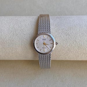 May include: A silver-tone Seiko Excaline quartz watch featuring a round face with a textured light dial, thin gold-tone hands, and a woven metal mesh bracelet. The watch face displays the Seiko logo and the word Excaline in black text.