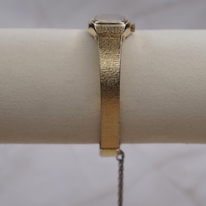 May include: A gold-toned wristwatch with a rectangular face and a textured band. The watch features a small, square watch face and a delicate chain attached to the band. The band has a woven texture.