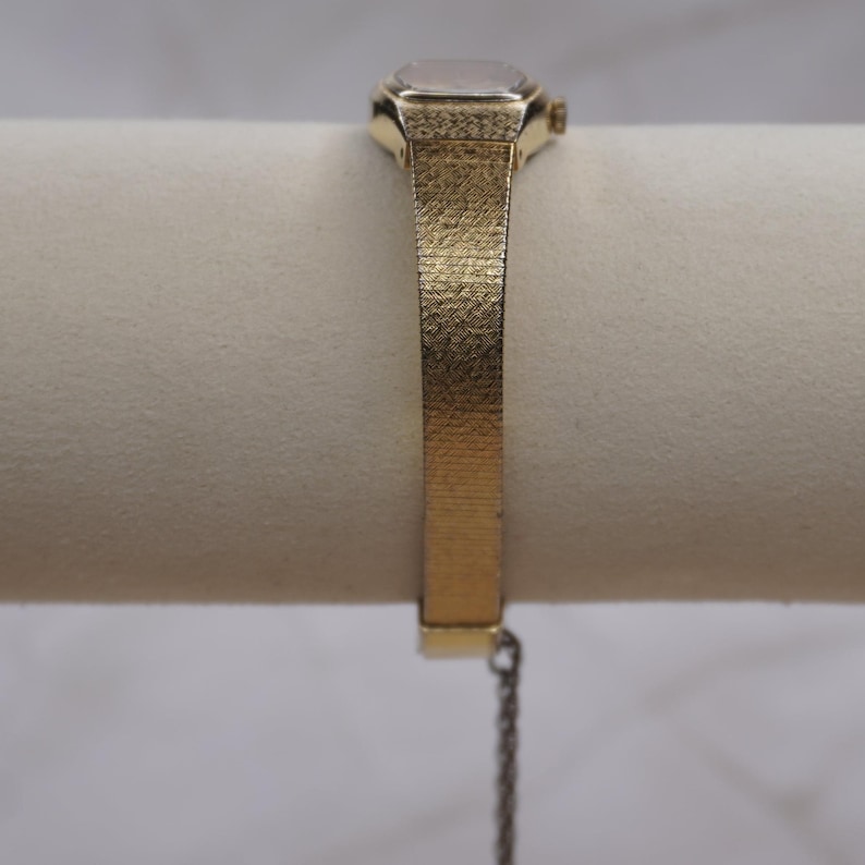 May include: A gold-toned wristwatch with a rectangular face and a textured band. The watch features a small, square watch face and a delicate chain attached to the band. The band has a woven texture.