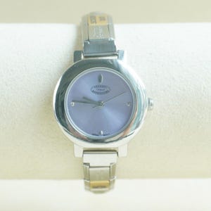 May include: A silver-tone wristwatch featuring a round case and a light purple dial. The dial displays the text Charmed Italy Expressions and Japan Movt. The watch has a metallic link bracelet with a silver finish and a small rectangular tag.